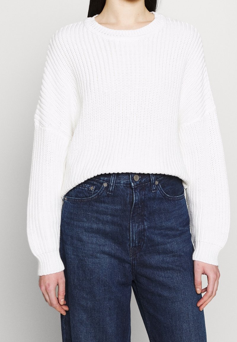 Personne portant un pull blanc à manches longues côtelé, rentré dans un jean taille haute bleu foncé, sur un fond uni.