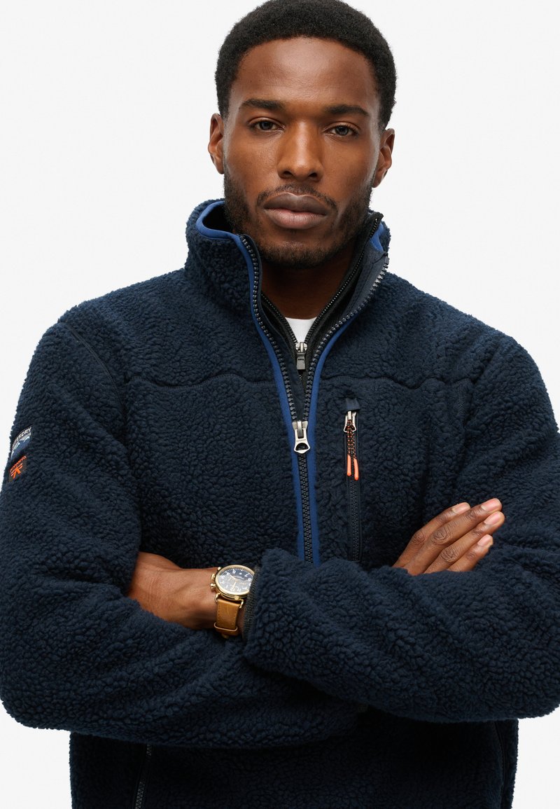Marineblauwe fleecejack met een gestructureerd oppervlak, met een hoge kraag, voorluiting met rits en een contrasterende oranje ritssluiting. Draag het met een horloge.