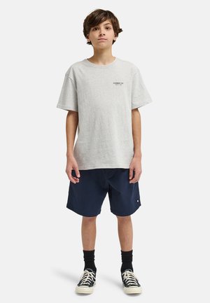 Chłopiec stojący przodem, ubrany w jasnoszary t-shirt, granatowe spodenki, czarne skarpetki i czarno-białe trampki na białym tle.