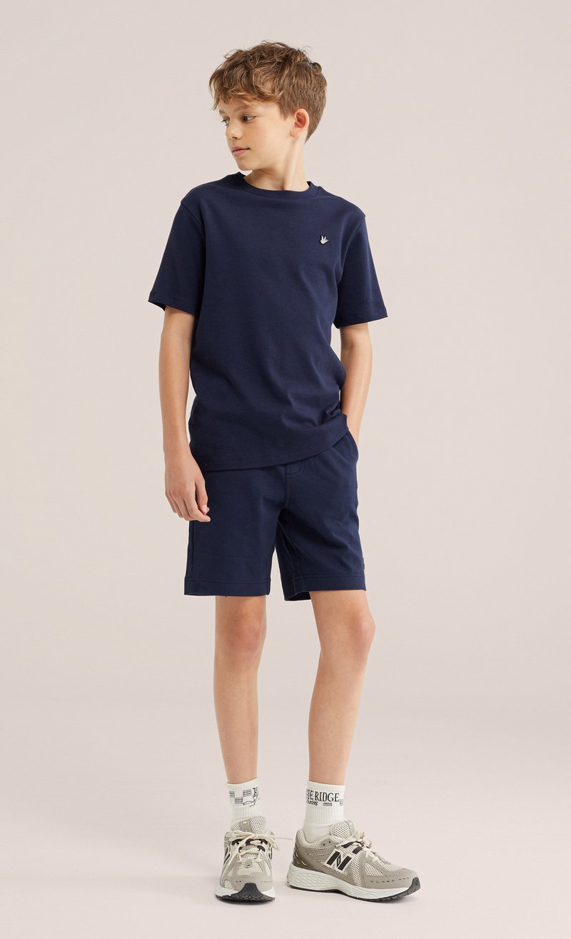 Garçon portant un T-shirt bleu marine et un short, des chaussettes blanches de marque et des baskets beiges, debout les mains dans les poches, regardant à gauche.