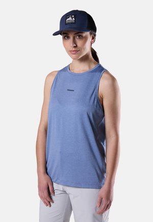 Woman wearing a blue sleeveless Rossignol top, white pants, and a navy Rossignol cap, standing against a plain light background.