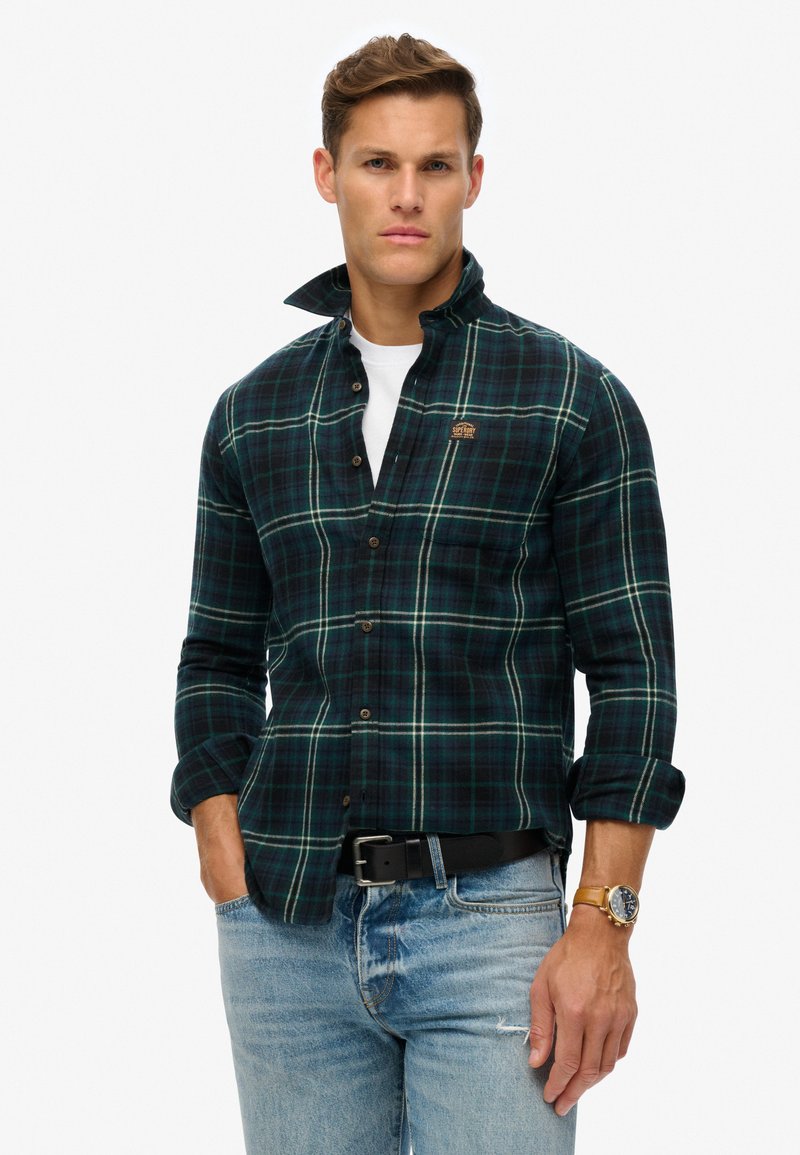 Donkergroen en zwart geruite flanellen shirt, wit T-shirt eronder, dichtgeknoopte kraag, opgerolde mouwen, gecombineerd met blauwe spijkerbroek en een zwarte riem.