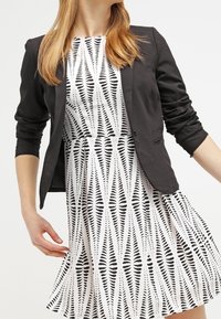 Musta istuva bleiseri yhdistettynä valkoiseen mekkoon, jossa rohkea musta lehtikuvio. Mekossa on levenevä helma ja lyhyet hihat.