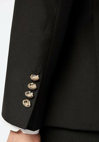 Manche de veste noire en tissu taillé, boutons en or avec un design complexe, et poignet de chemise blanche dépassant en dessous.