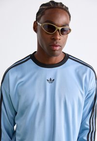 Light blue long-sleeve athletic shirt with black stripe accents, featuring an Adidas logo on the chest. Pair of light-colored sunglasses.