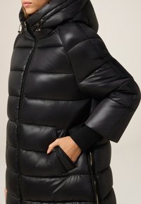 Giacca nera imbottita con finitura lucida, dotata di cappuccio, tasche con zip e polsini a coste per maggiore calore e comfort.