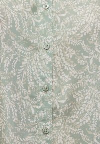 Tissu vert clair à motif feuillu blanc avec cinq petits boutons cousus en ligne verticale au centre.