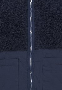 Veste bleu marine devant montrant un haut en polaire texturé, une partie inférieure en tissu matelassé, deux poches basses et une fermeture éclair centrale argentée.