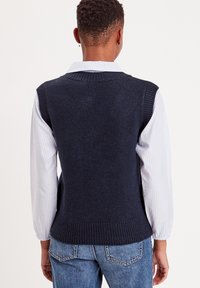 Marineblå strikket uden ærmer sweater med ribbestrikket nederste kant, båret over en hvid og blå stribet langærmet skjorte, med en afslappet pasform og struktur.