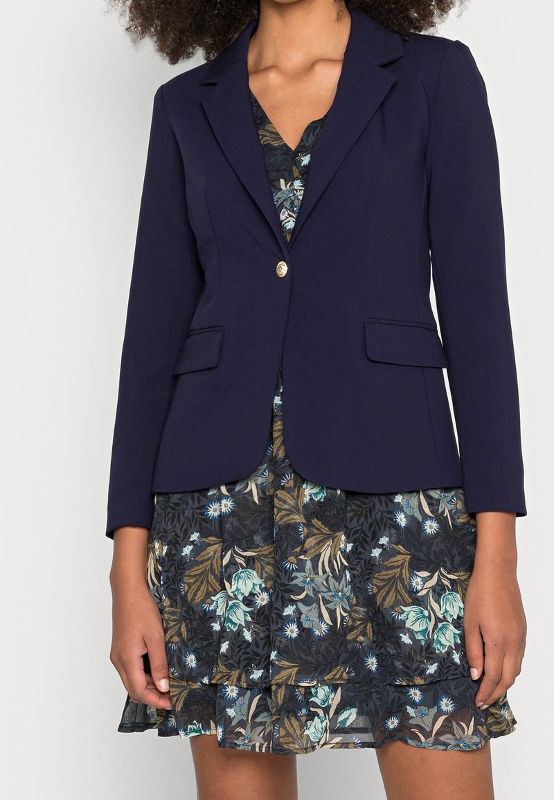 Blazer blu navy con due tasche frontali e un singolo bottone dorato, indossato sopra un vestito a fantasia floreale con accenti blu e verdi.