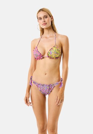 Donna che indossa un bikini con motivo floreale nei colori rosa, giallo e viola, in piedi davanti a uno sfondo grigio chiaro uniforme.