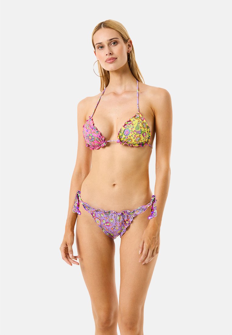 Mujer con bikini de estampado floral en colores rosa, amarillo y púrpura, de pie frente a un fondo gris claro liso.