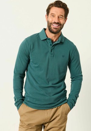 Mann mit Bart, der ein petrolfarbenes Langarm-Poloshirt und beige Hose trägt, lächelt mit den Händen in den Taschen vor einfarbigem Hintergrund.