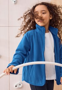 Jachetă albastră din fleece cu guler înalt, fermoar frontal și buzunare laterale, purtată peste o cămașă albă, cu părul creț care acoperă parțial fața.