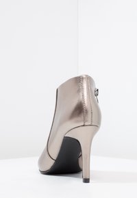 Zapato de tacón alto en bronce metálico con un diseño elegante, punta afilada, textura suave y un detalle de cremallera en la parte trasera. Suela negra.