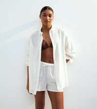 Vit linneknappskjorta med uppvikta ärmar, parat med ljusa shorts. Brun baddräktstopp synlig under. Slät textur, avslappnad passform.