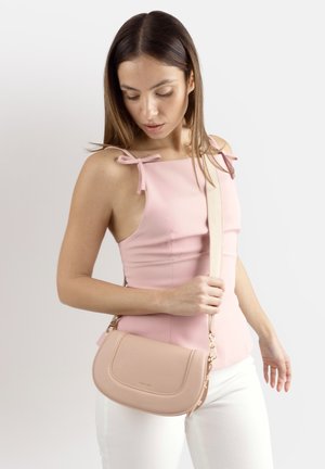 Donna con un top rosa senza maniche e pantaloni bianchi tiene una borsa a tracolla beige con una cinghia larga su uno sfondo semplice.