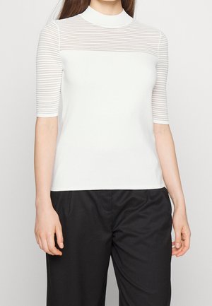 Žena v bílém krátkém tričku s průsvitným průkrčníkem a rukávy s vodorovnými pruhy, v kombinaci s volnými černými kalhotami.