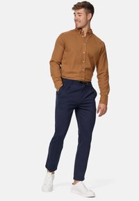 Camisa de botones marrón con cuello puntiagudo, combinada con pantalones azul marino a medida y zapatillas blancas, creando un aspecto casual pero pulido.