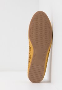 Semelle d’une chaussure plate jaune avec motif adhérent texturé et motif floral perforé sur les côtés, positionnée verticalement sur fond blanc.