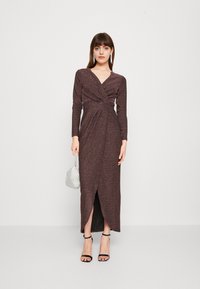 Robe longue marron foncé avec des manches longues, un col en V et un design enroulé, comportant une fente à l'avant. Portée avec des sandales noires à brides et un sac argenté.