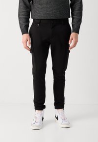 Svarte cargo bukser med tapered fit, med sidelommer og glatt tekstur, kombinert med hvite høye sneakers.
