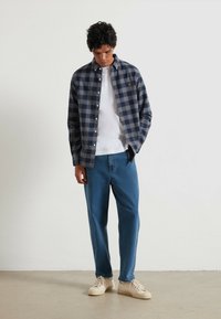 Chemise à carreaux pour homme en bleu marine et gris, design à boutons, portée sur un t-shirt blanc, associée à un jean bleu et des baskets beiges.