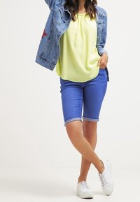 Veste en denim avec détail étoile rouge sur un chemisier jaune vif, associée à un short en denim bleu et des baskets blanches. Combinaison de tenue décontractée.