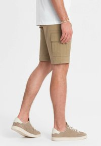 Pantaloni cargo beige con due tasche laterali, realizzati in tessuto morbido. Abbinati a sneakers in suede color cuoio con suola bianca e dettagli in bianco.