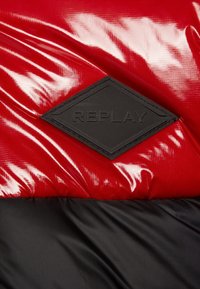 Sort diamantformet "REPLAY" lap på skinnende rød og mat sort quiltet stof, sandsynligvis del af en jakke.