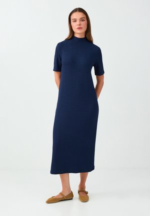 Femme debout, les mains derrière le dos, portant une robe midi bleu marine à manches courtes et des chaussures plates marron sur un fond clair uni.