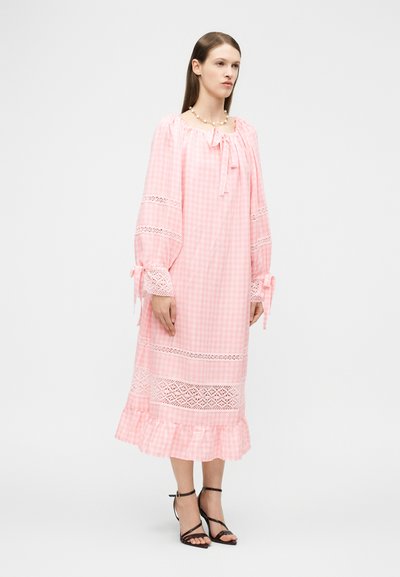 Γυναίκα που φοράει μακρύ ροζ φόρεμα γκίνχαμ με δαντέλες, λεπτομέρειες με φιόγκους στα μανίκια και το ντεκολτέ, και μαύρα σανδάλια με λουράκια και τακούνι.