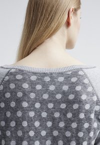 Suéter gris con un patrón de puntos en gris claro, con cuello redondo y mangas raglán. Textura suave con un escote liso en contraste.