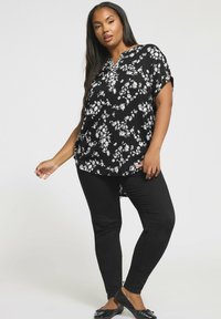 Kvinna i en svart och vit blommig topp, svarta figurnära byxor och svarta ballerina skor, som står mot en enkel ljus bakgrund.