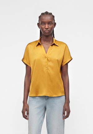 Sötét bőrű nő, fonott hajjal, mustár sárga rövid ujjú gombos ingben és világoskék farmernadrágban, szemben áll.