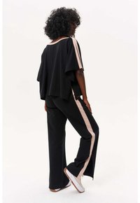 Ensemble noir décontracté, haut et pantalon avec des bandes latérales rose clair. Le tissu semble doux ; pantalon évasé, baskets visibles, design simple.