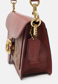 Ruskea nahka- ja kangas Coach -laukkua, jossa on kultaiset yksityiskohdat ja signatuurilogo-tagi, joka roikkuu metalliketjusta.