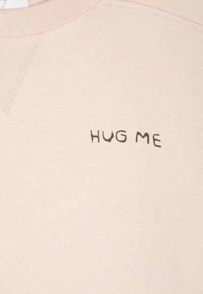 Tissu beige en gros plan avec texte noir "HUG ME" imprimé près du centre supérieur sur un vêtement à col côtelé.