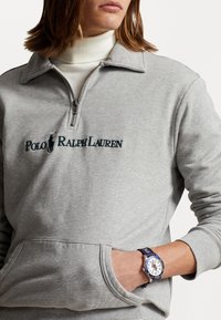 Šedá mikina s čtvrtinovým zipem a vyšitým logem "Polo Ralph Lauren". Obsahuje přední kapsu a model nosící námořnické hodinky.