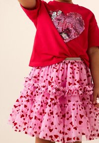 Enfant portant un t-shirt rouge avec un grand cœur rose en sequins et une jupe rose à volants couverte de motifs de cœurs rouges.