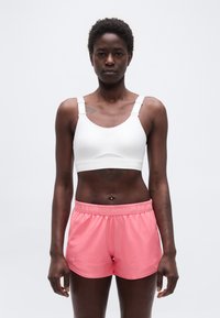 Γυναίκα αθλήτρια που στέκεται με το πρόσωπο προς τα εμπρός, φορώντας λευκό αθλητικό σουτιέν και ροζ αθλητικά σορτς Under Armour, σε ανοιχτό φόντο.