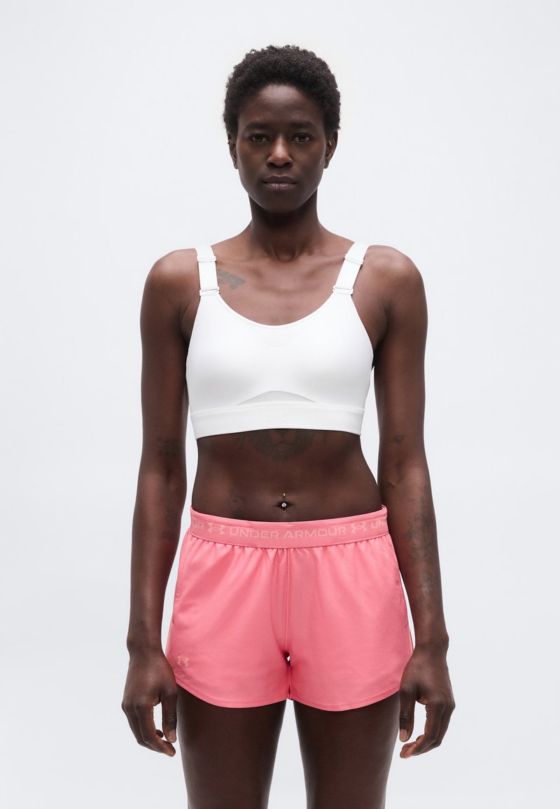 Γυναίκα αθλήτρια που στέκεται με το πρόσωπο προς τα εμπρός, φορώντας λευκό α θλητικό σουτιέν και ροζ αθλητικά σορτς Under Armour, σε ανοιχτό φόντο.