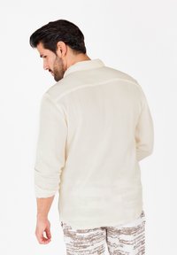 Camisa de manga larga de color beige claro y texturizada, con las mangas remangadas, que presenta un cuello clásico y costuras horizontales sutiles en la parte trasera.