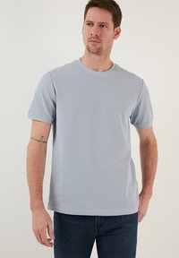 Tricou bleu deschis cu guler rotund, dintr-un material moale și texturat. Mâneci scurte cu o croială lejeră, purtat cu blugi bleu închis.