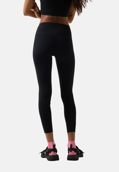 Frau in schwarzen Leggings, schwarzem kurzer Oberteil, pinkfarbenen Socken und schwarzen Sandalen, stehend mit verschränkten Armen, von hinten vor einem weißen Hintergrund gesehen.