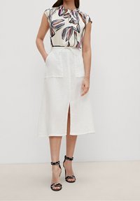 Blusa de crema con estampado floral y mangas cortas, combinada con una falda de lino blanca hasta la rodilla con bolsillos frontales y una abertura. Tacones negros con tiras.