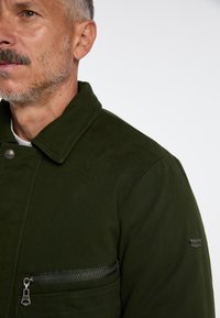 Olivengrøn jakke lavet af glat stof med spids krave, enkelt brystlomme med lynlås og mærke på ærmet.