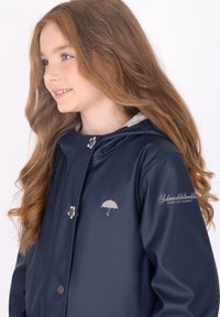 Tamsiai mėlynas vandeniui atsparus švarkas su sidabro akcentais, užsegamas spaustukais ir turintis skėčio logotipą. Savybė - gobtuvas ir minkštas pilkas pamušalas.