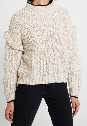 Beige grofgebreide trui met ruchtdetails op de mouwen, gecombineerd met zwarte spijkerbroek bij een staand persoon tegen een effen achtergrond.