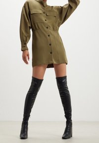 Persona che indossa un mini abito verde oliva con bottoni, maniche lunghe e stivali neri con tacco sopra il ginocchio, in piedi contro uno sfondo neutro.